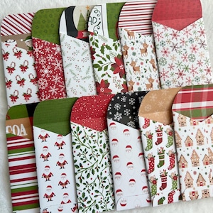 May include: A set of 14 Christmas-themed gift card holders. The holders are made of paper and feature various holiday designs, including Santa Claus, reindeer, snowflakes, and gingerbread houses.