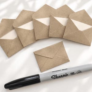 May include: A set of six small brown kraft paper envelopes, five of which are open and one is closed, arranged with a grey Sharpie fine point permanent marker positioned horizontally below them on a white surface.