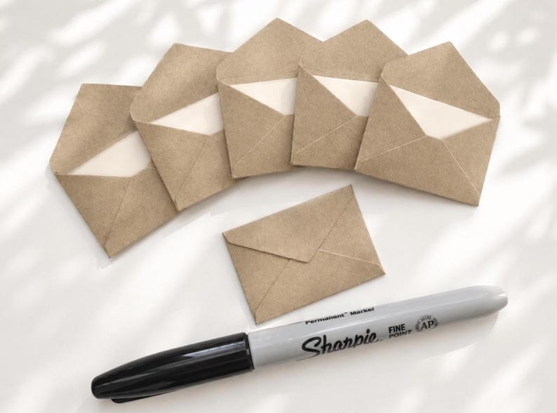 May include: A set of six small brown kraft paper envelopes, five of which are open and one is closed, arranged with a grey Sharpie fine point permanent marker positioned horizontally below them on a white surface.