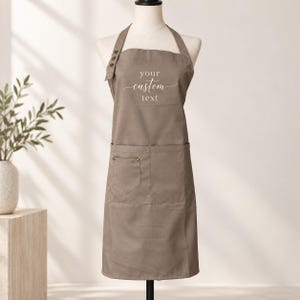May include: A taupe apron featuring a neck strap with three decorative buttons on the right side. The chest area displays the white text "your custom text" in a script and sans-serif font. The apron includes a large front pocket with a smaller section.