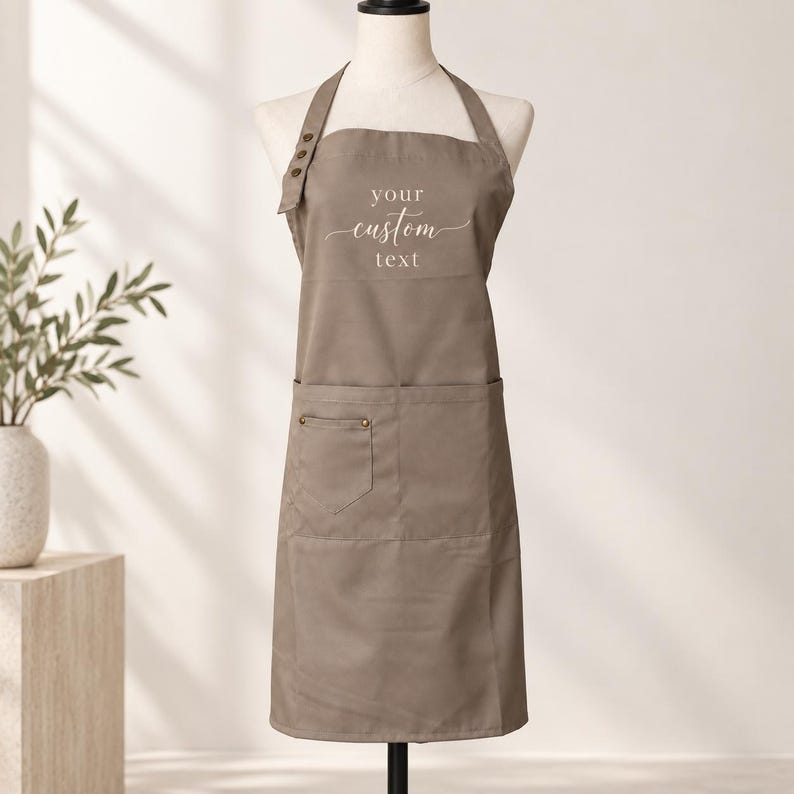 May include: A taupe apron featuring a neck strap with three decorative buttons on the right side. The chest area displays the white text "your custom text" in a script and sans-serif font. The apron includes a large front pocket with a smaller section.