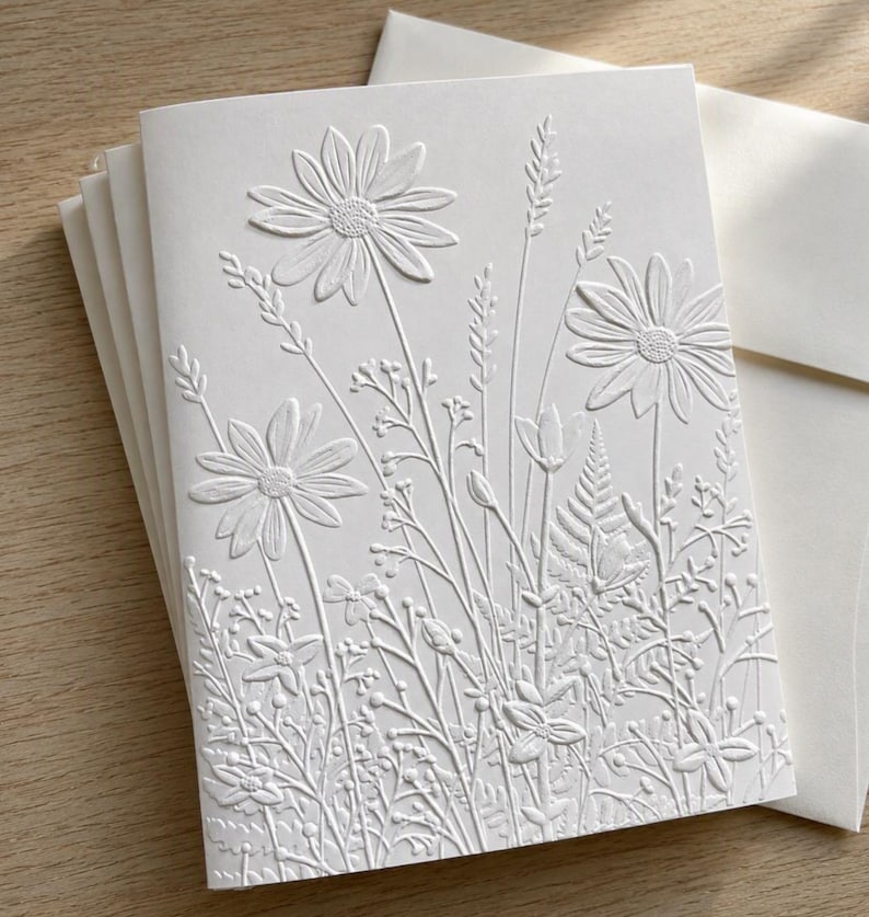 Puede incluir: Una tarjeta de felicitaci&oacute;n blanca con un dise&ntilde;o en relieve de varias flores silvestres y hierbas. La textura crea un patr&oacute;n floral monocrom&aacute;tico con margaritas, helechos y tallos delicados en la parte frontal de la tarjeta de papel plegada.
