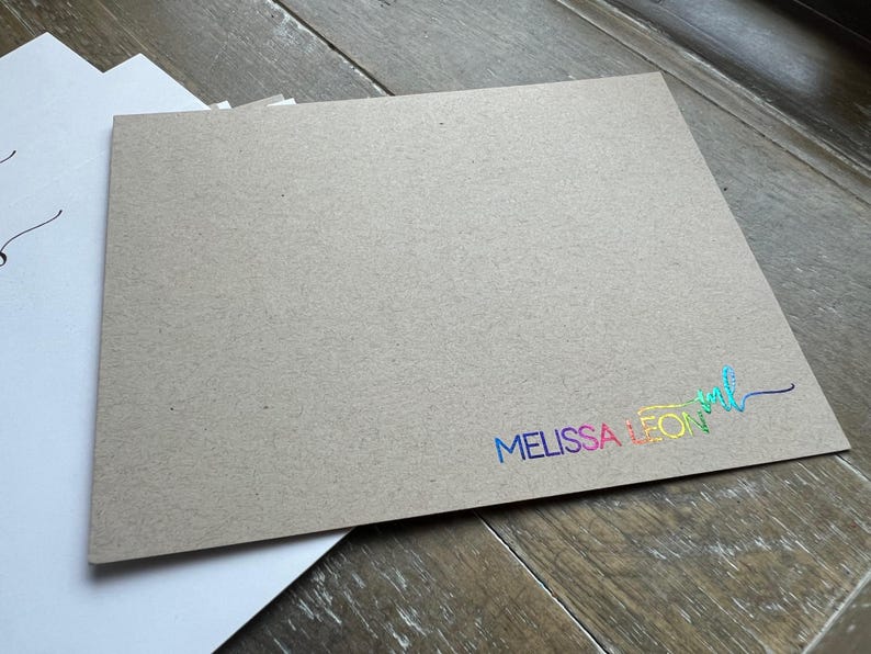 May include: A rectangular piece of tan kraft paper cardstock featuring the text MELISSA LEON in a vibrant, iridescent rainbow foil finish. The text is positioned in the bottom right corner, accompanied by a stylized script monogram of the letters ml.