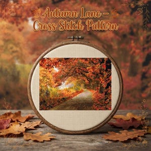 May include: A circular wooden embroidery hoop containing a cross stitch pattern of an autumn forest path with vibrant orange, red, and yellow foliage. The text "Autumn Lane - Cross Stitch Pattern" is printed in a stylized font above the hoop.