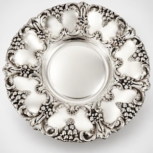 May include: A round silver plate featuring a smooth, polished center basin surrounded by an ornate, raised border. The rim is decorated with intricate, embossed grape cluster motifs and scrolling leaf patterns in a metallic finish.