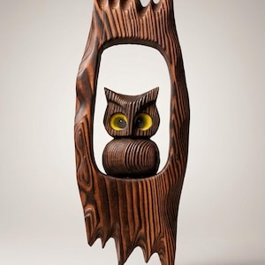 May include: A dark brown carved wooden sculpture of an owl with bright yellow eyes, positioned within a vertical, textured wooden frame that mimics the appearance of a hollowed-out tree trunk with jagged, pointed edges at the top and bottom.