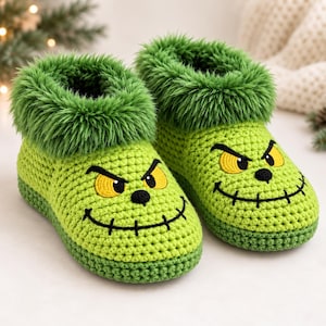 Op de afbeelding: Een paar limoengroene gehaakte pantoffels met een pluizige groene rand van imitatiebont. De pantoffels hebben een geborduurd gezicht met gele ogen, zwarte wenkbrauwen en een brede, gestikte zwarte grijns op de voorzijde.