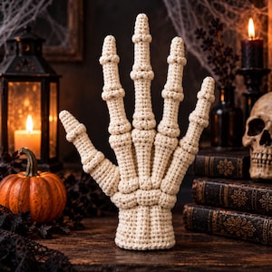 Puede incluir: Una escultura de mano de esqueleto tejida a ganchillo en color crema, con articulaciones de dedos detalladas y una base de muñeca cilíndrica. La mano está elaborada con puntadas de hilo visibles, creando una textura tridimensional.