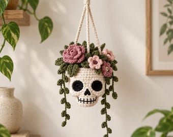 May include: A crocheted skull-shaped hanging planter in off-white yarn with black eye sockets and stitched teeth. The skull is topped with various crocheted flowers in shades of pink and dark green leaves, with long trailing green vines hanging down.