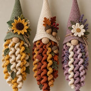 Könnte beinhalten: Drei hängende Häkel-Gnom-Ornamente mit spitzen Hüten. Der erste hat einen grünen Hut mit Sonnenblume, der zweite einen cremefarbenen Hut mit Eicheln und Blättern, und der dritte einen lavendelfarbenen Hut mit weißer Blume und lila Zweigen.