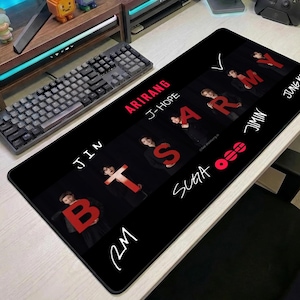 May include: A black rectangular desk mat featuring seven individual portraits of people, each holding a large red letter. The letters spell out BTS ARMY. The text ARIRANG, JIN, J-HOPE, SUGA, JIMIN, JUNGKOOK, and RM is printed in white on the mat.