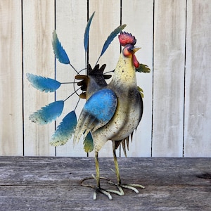 May include: A metal rooster garden sculpture featuring a cream-colored body with distressed brown speckles, a bright red comb and wattle, and a large, circular tail fan composed of individual blue, feather-shaped metal blades attached to wire spokes.