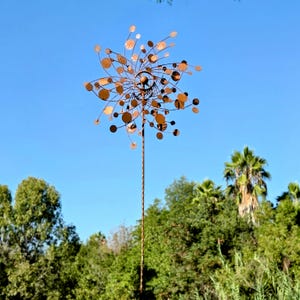 May include: A copper-colored metal wind spinner featuring a central hub with multiple radiating wire arms, each tipped with small circular discs. The structure is mounted on a tall, thin, twisted metal pole designed for outdoor garden display.