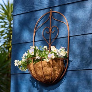 May include: A wall-mounted planter featuring a rust-colored metal frame with an arched top and a heart-shaped design element. The planter contains a brown coconut fiber liner filled with white and light purple pansy flowers.