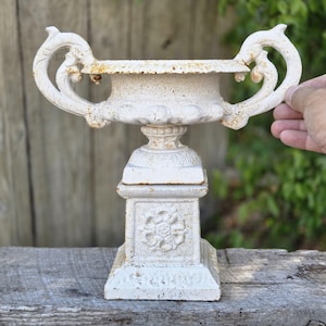 May include: A white cast iron urn planter featuring two ornate, curved handles on the sides and a square base decorated with a raised floral motif. The surface shows signs of weathered paint and rust, revealing the texture of the metal material.