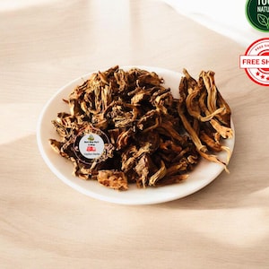 Puede incluir: Un plato blanco lleno de un montón de trozos de plantas secas, marrones y fibrosas. La textura es rugosa e irregular, con algunas piezas que parecen curvadas o retorcidas. Un pequeño logotipo circular con texto es visible en el montón.
