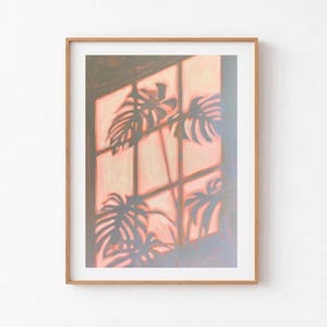Puede incluir: Una lámina de arte enmarcada que muestra la pintura de una ventana con marco de cuadrícula. Los cristales están llenos de una suave luz melocotón y rosa, mientras que sombras gris oscuro de hojas de monstera se proyectan.