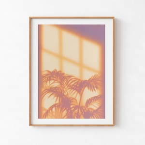 Puede incluir: Una lámina de arte enmarcada que presenta una imagen de tonos cálidos y enfoque suave de una cuadrícula de ventana que proyecta luz y sombras sobre la silueta de una palmera. La obra está en un marco de madera clara con un paspartú blanco.