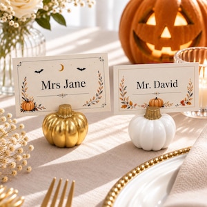 May include: Two rectangular place cards with black text reading Mrs Jane and Mr. David, decorated with autumn leaves, bats, and a crescent moon. Each card is held by a small pumpkin-shaped stand, one in metallic gold and one in white.