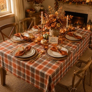 May include: A dining table set for autumn with a plaid tablecloth in orange, brown, and white. The table features white plates, orange napkins with decorative rings, amber glassware, and a centerpiece of pumpkins and fall foliage with a Give Thanks sign.