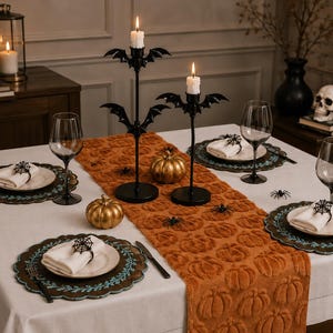 May include: A dining table setting featuring a white tablecloth, an orange pumpkin-patterned table runner, and four place settings with white plates, white napkins, and black spiderweb napkin rings. Two black bat-themed candle holders hold lit candles.