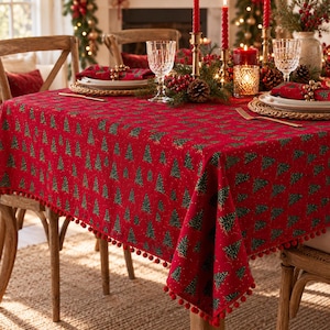 May include: A rectangular red tablecloth featuring a repeating pattern of green Christmas trees and small white dots, finished with a decorative red pom-pom trim along the edges. The fabric is draped over a dining table set for a holiday meal.