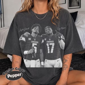 May include: A charcoal grey short-sleeve t-shirt featuring a black and white graphic print of football players Jayden Daniels and Terry McLaurin in Washington Commanders jerseys with the numbers 5 and 17, including their names printed above them.