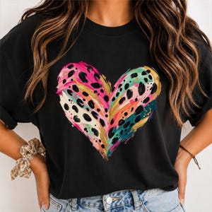 May include: A black t-shirt featuring a large, central heart graphic. The heart is composed of vibrant, abstract brushstrokes in shades of hot pink, teal, and gold, overlaid with a black leopard print pattern that creates a textured, artistic effect.