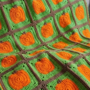 May include: A crochet blanket composed of square motifs, each featuring a bright orange pumpkin centered on a vibrant green background. The squares are joined together with a dark brown yarn border, creating a grid pattern across the textile.