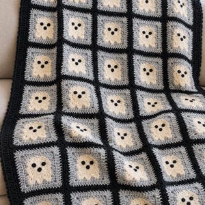May include: A crochet blanket featuring a repeating pattern of square motifs. Each square contains a cream-colored ghost figure with two black eyes and a black mouth, set against a light gray background, all bordered by thick black yarn lines.
