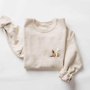 May include: A beige crewneck sweatshirt featuring a small, embroidered graphic on the left chest of a brown and cream-colored creature lying down with its eyes closed and three small white circles floating above its head.