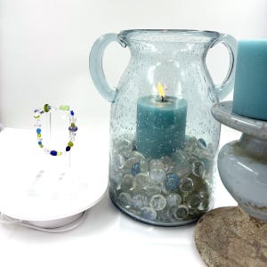 May include: A beaded bracelet with clear, blue, and green glass beads on a stand, next to a light blue glass vase containing a lit teal candle and clear glass pebbles, with a teal pillar candle on a rustic pedestal base nearby.