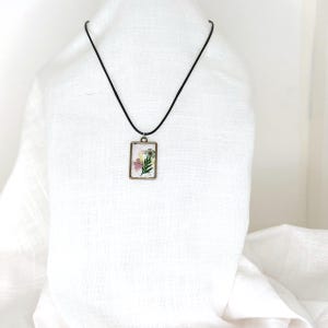 May include: A rectangular pendant necklace featuring a bronze-toned metal frame containing pressed flowers in shades of pink, yellow, and green. The pendant hangs from a simple black cord, creating a clear contrast against the light-colored fabric.
