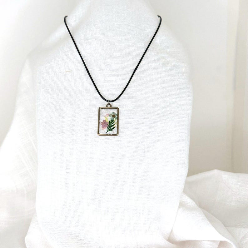 May include: A rectangular pendant necklace featuring a bronze-toned metal frame containing pressed flowers in shades of pink, yellow, and green. The pendant hangs from a simple black cord, creating a clear contrast against the light-colored fabric.