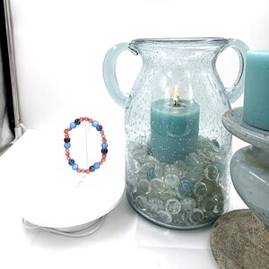 May include: A circular beaded bracelet featuring alternating light blue, dark purple, and faceted copper-toned beads. The bracelet is displayed on a white stand next to a large, light blue glass vase filled with clear glass pebbles and a teal candle.