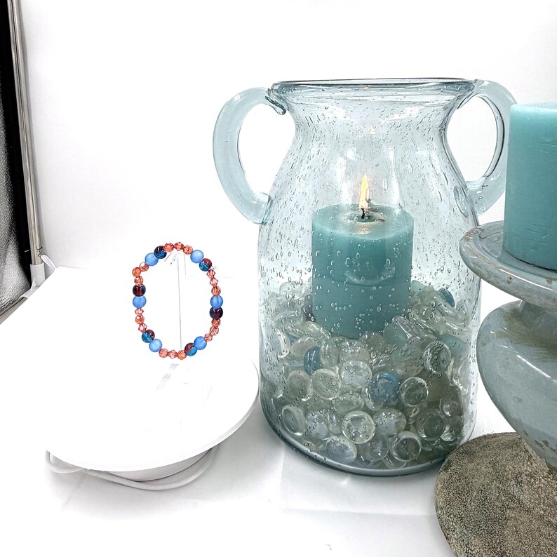 May include: A circular beaded bracelet featuring alternating light blue, dark purple, and faceted copper-toned beads. The bracelet is displayed on a white stand next to a large, light blue glass vase filled with clear glass pebbles and a teal candle.
