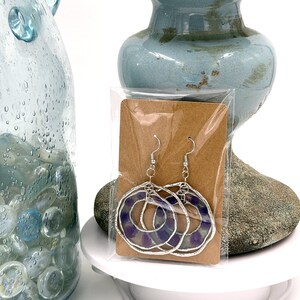 May include: A pair of silver-toned dangle earrings featuring circular frames with wavy edges. Each earring contains a translucent purple circular insert with a marbled pattern. The earrings are attached to a brown rectangular display card in plastic.