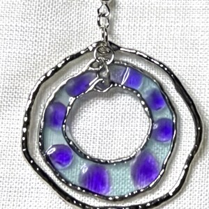 May include: A silver-toned metal pendant featuring two concentric circular frames. The inner circle contains a translucent light blue resin or glass insert decorated with distinct, rounded purple spots. A silver chain link connects the two circles.