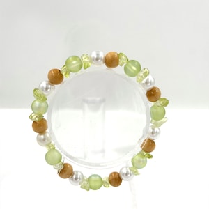 May include: A beaded bracelet featuring a repeating pattern of smooth white pearls, light green frosted glass beads, polished brown wooden spheres, and small, irregular light green stone chips arranged in a circular design on a clear stand.