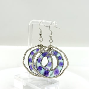 May include: A pair of dangling earrings featuring circular frames with a textured silver-toned metal edge. Each earring contains a translucent light blue resin center embedded with small, rounded purple accents, suspended from silver-toned metal hooks.