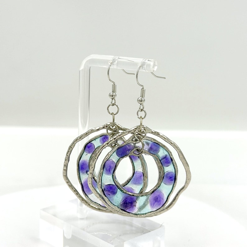 May include: A pair of dangling earrings featuring circular frames with a textured silver-toned metal edge. Each earring contains a translucent light blue resin center embedded with small, rounded purple accents, suspended from silver-toned metal hooks.