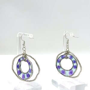 May include: A pair of dangle earrings featuring two nested circular frames. The outer frame is a textured silver-tone metal ring, while the inner frame is a translucent light blue glass ring with purple circular spots, suspended by a silver chain.