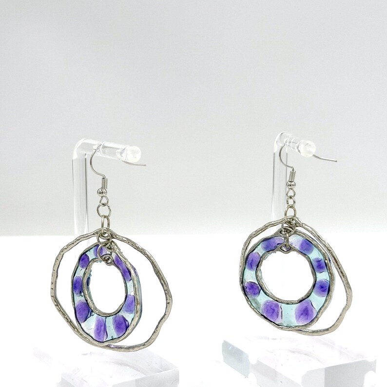 May include: A pair of dangle earrings featuring two nested circular frames. The outer frame is a textured silver-tone metal ring, while the inner frame is a translucent light blue glass ring with purple circular spots, suspended by a silver chain.