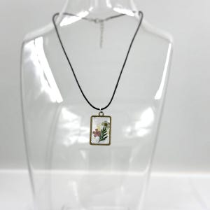 May include: A rectangular pendant necklace featuring a bronze-toned metal frame enclosing a clear resin insert with embedded pressed flowers, including a pink blossom and green leaves, suspended from a simple black cord with a metal clasp.