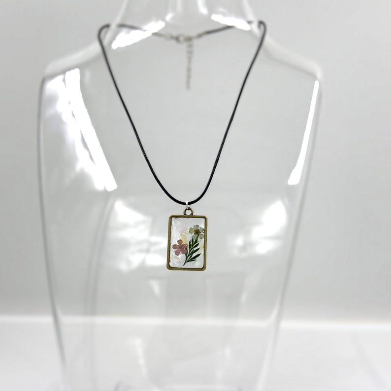 May include: A rectangular pendant necklace featuring a bronze-toned metal frame enclosing a clear resin insert with embedded pressed flowers, including a pink blossom and green leaves, suspended from a simple black cord with a metal clasp.