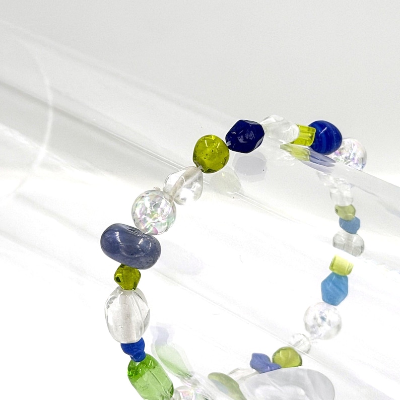 May include: A beaded bracelet featuring a variety of shapes and sizes of beads in shades of clear, lime green, royal blue, and periwinkle. The beads are strung on a clear cord and include faceted, smooth, and iridescent finishes throughout the design.