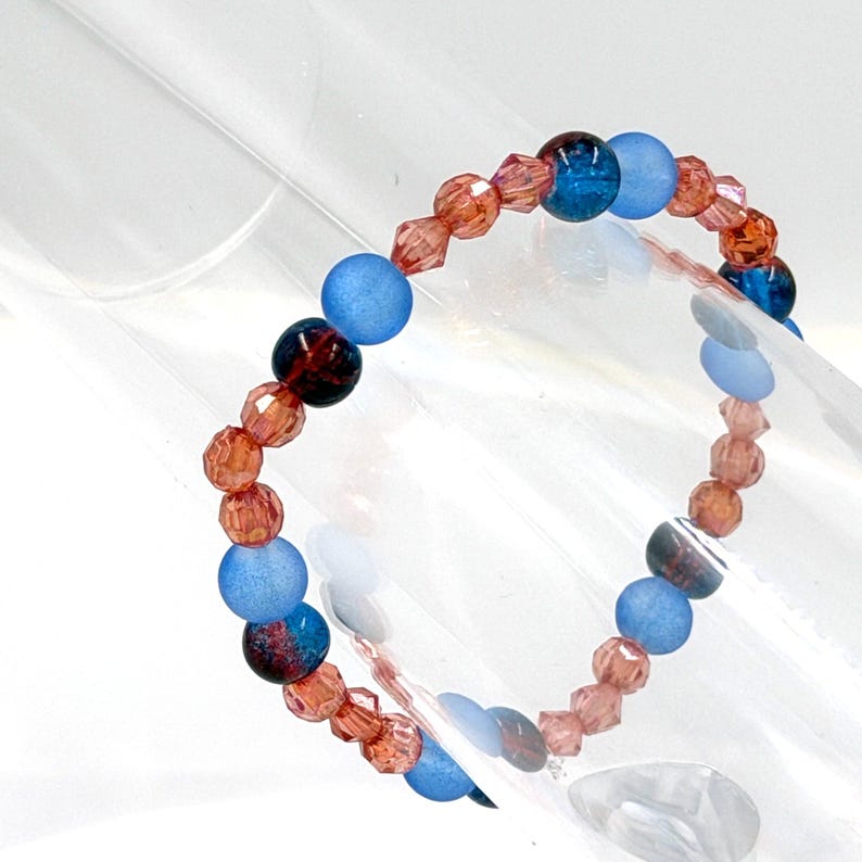 May include: A beaded bracelet featuring a repeating pattern of faceted copper-colored crystal beads, smooth matte light blue round beads, and dark blue marbled round beads. The beads are strung together in a circular arrangement on a flexible cord.