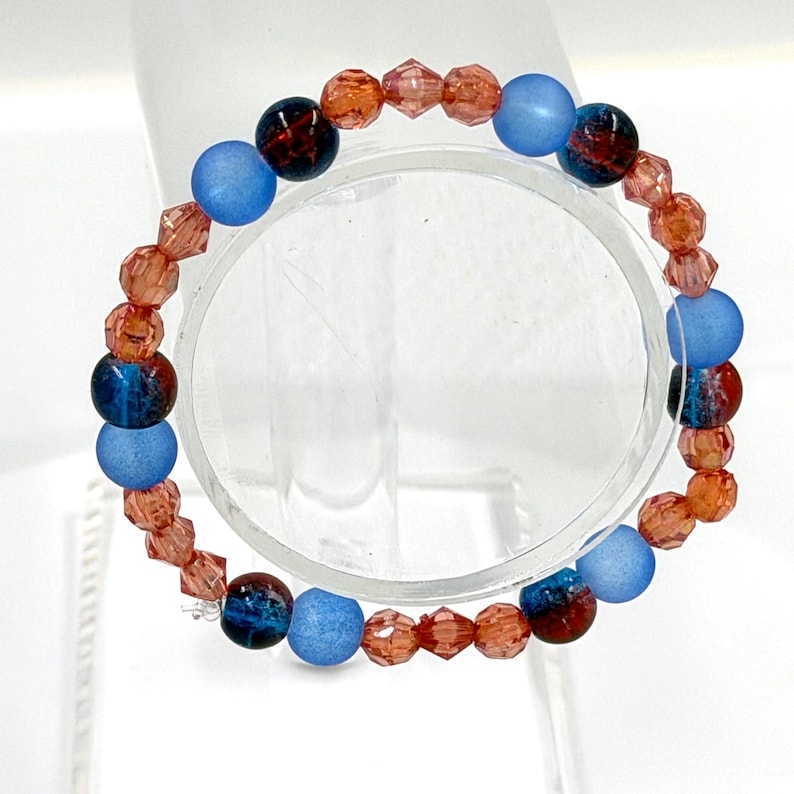 May include: A circular bracelet composed of alternating beads, featuring matte light blue spheres, dark blue marbled spheres, and faceted translucent orange crystals arranged in a repeating pattern around the entire circumference of the jewelry piece.