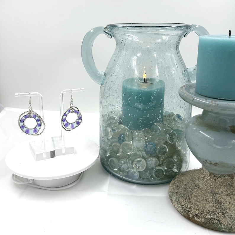 May include: A pair of silver-toned hoop earrings with purple and white circular patterns, displayed on clear stands. Beside them sits a light blue glass pitcher filled with clear glass pebbles and a lit teal candle, plus a separate teal pillar candle.