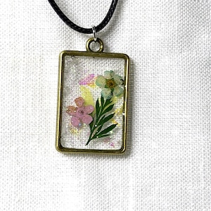 May include: A rectangular pendant necklace featuring a bronze-toned metal frame containing clear resin embedded with two small pressed flowers, one pink and one light blue, along with green leaves and fine iridescent glitter particles.
